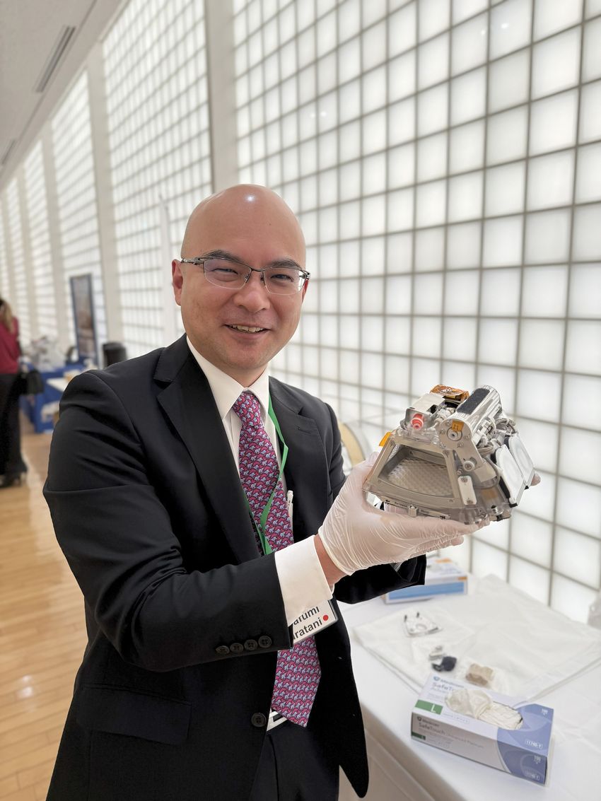 Photograph of Masafumi Muratani, a molecular biologist at the University of Tsukuba, holding a model of a mouse habitat cage unit. Muratani is wearing a white collared shirt, black suit jacket and pants, a red patterned tie, and white nitrile gloves.