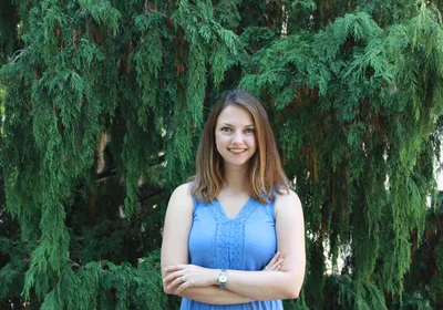 Image of Niki Spahich. She smiles at the camera and wears a blue top. She stands with her arms crossed in front of a green background.