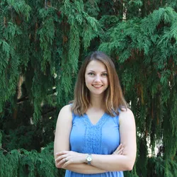 Image of Niki Spahich. She smiles at the camera and wears a blue top. She stands with her arms crossed in front of a green background.