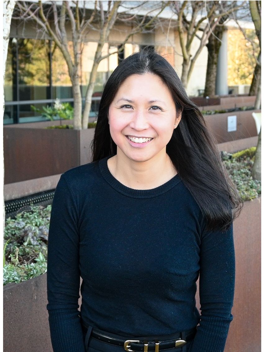 Jamie Lo, a maternal-fetal medicine specialist at the Oregon Health and Science University, stands outside in front of a row of planters with trees in them. She has long, straight dark hair and is wearing a navy, long-sleeved top.