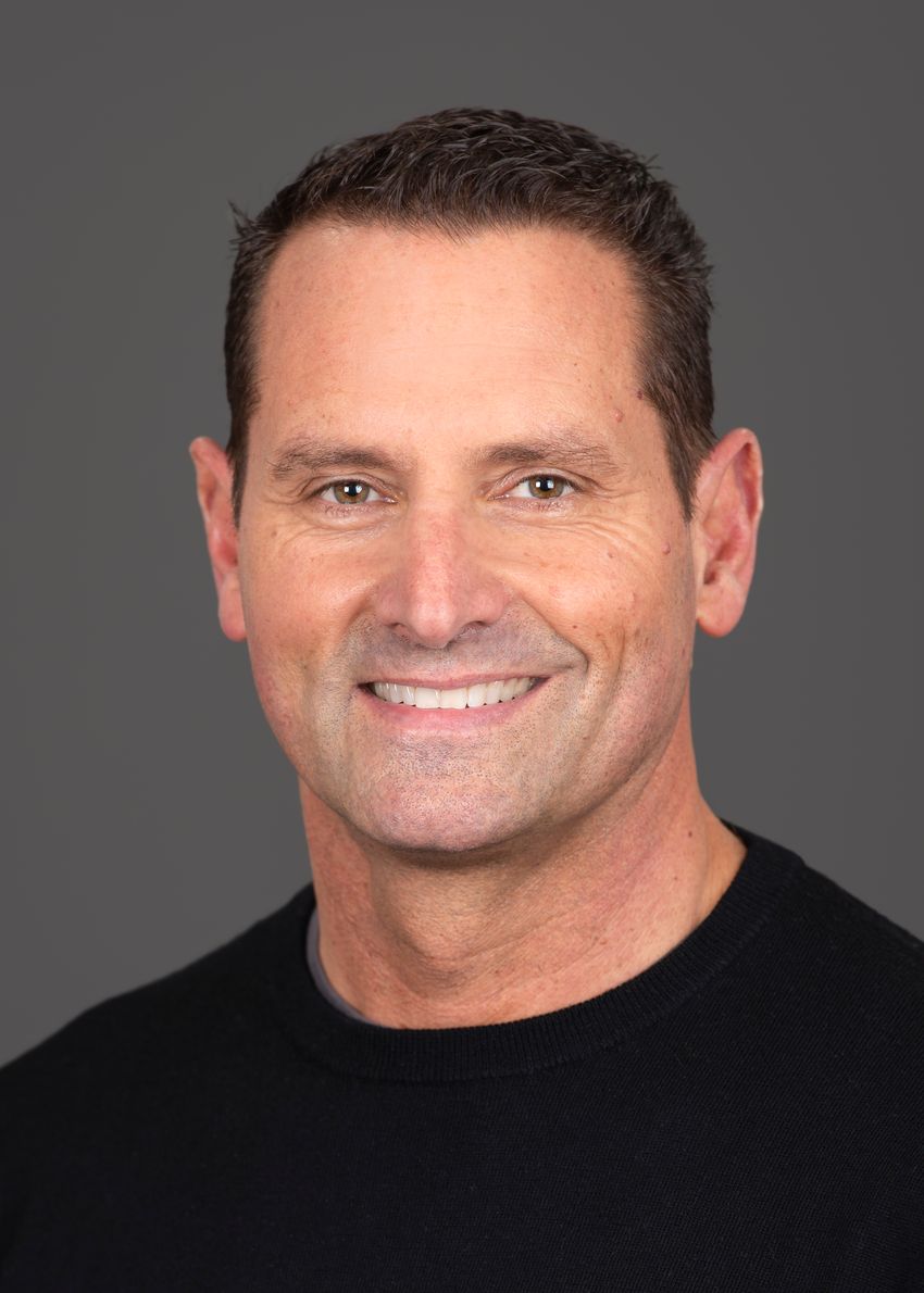 Brian Scottoline, a neonatologist at the Oregon Health and Science University, is smiling at the camera in a professional photo with a grey background. He has short brown hair and is wearing a black shirt.