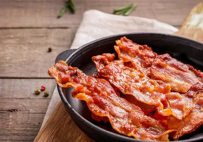 Hot fried crispy bacon slices in skillet on wooden background. Fat-associated sensory cues such as bacon odors in maternal diet may influence offsprings’ predisposition to obesity during adulthood. 