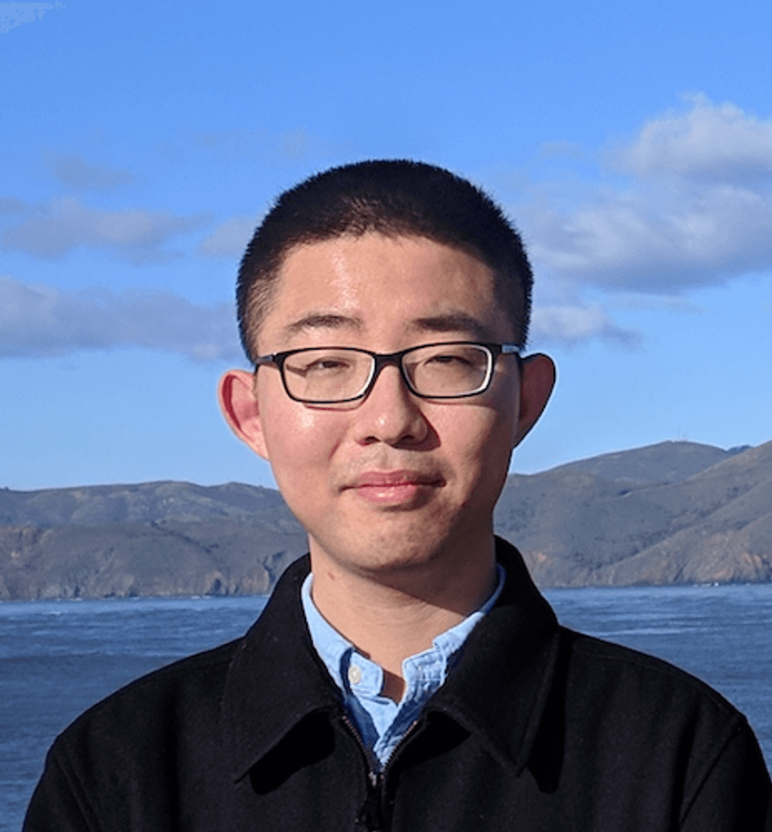 Photograph of Li Wang, a postdoctoral researcher in Arnold Kriegstein’s lab studying human brain development. Wang is standing in front of a background of mountains and blue water on a clear day. He has short dark hair and is wearing glasses and a black jacket over a blue shirt.