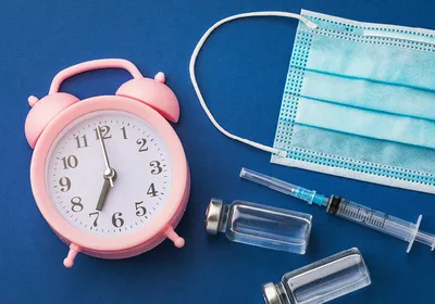 A pink alarm clock, a blue surgical mask, a syringe, and two vials lie on a blue surface, indicating how the time of cancer immunotherapy administration may influence their outcomes.
