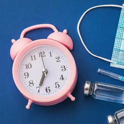 A pink alarm clock, a blue surgical mask, a syringe, and two vials lie on a blue surface, indicating how the time of cancer immunotherapy administration may influence their outcomes.