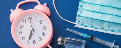 A pink alarm clock, a blue surgical mask, a syringe, and two vials lie on a blue surface, indicating how the time of cancer immunotherapy administration may influence their outcomes. 