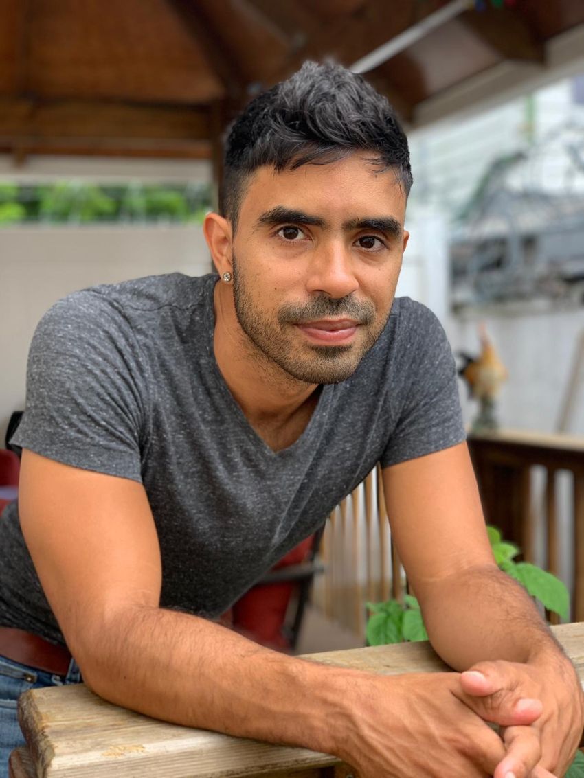 Jose Valdez, wearing a grey heathered T-shirt, stud earring, brown belt, and blue jeans, smiles at the camera.