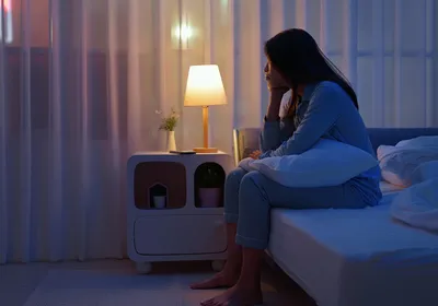 Image of a young woman sitting alone by the bedside. She appears troubled and unable to sleep.