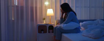 Image of a young woman sitting alone by the bedside. She appears troubled and unable to sleep.