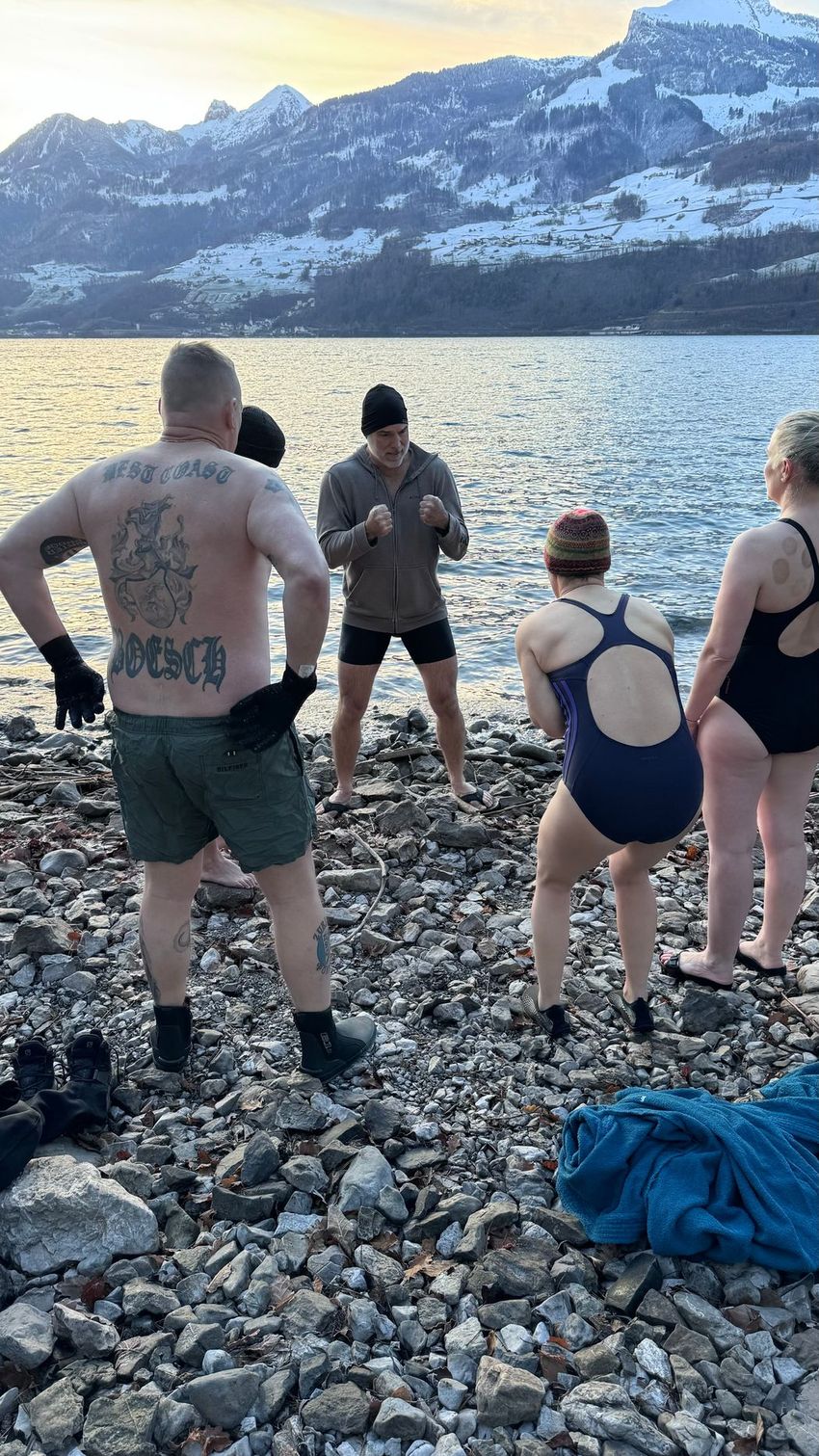 Image of François Haman speaking with people in swimsuits in front of a lake with snowy mountains in the background.