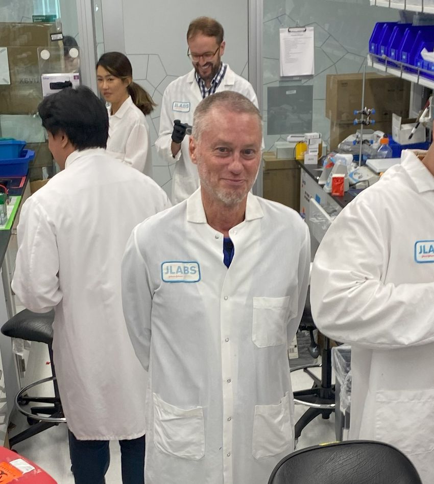 Timothy Friede stands in a white lab coat in the middle of a lab surrounded by scientists carrying out experiments. He smiles at the camera.