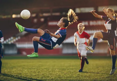 Three female soccer players watch as one of their teammates does a bicycle kick on the soccer pitch.