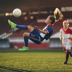 Three female soccer players watch as one of their teammates does a bicycle kick on the soccer pitch.