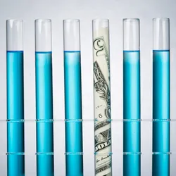 Photograph of six glass test tubes standing in a rack on a gray background. Five of the test tubes are filled with blue liquid, while the fourth from the left has a five-dollar bill in it to represent the cost of research. 