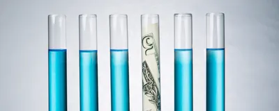 Photograph of six glass test tubes standing in a rack on a gray background. Five of the test tubes are filled with blue liquid, while the fourth from the left has a five-dollar bill in it to represent the cost of research. Photograph of six glass test tubes standing in a rack on a gray background. Five of the test tubes are filled with blue liquid, while the fourth from the left has a five-dollar bill in it to represent the cost of research.