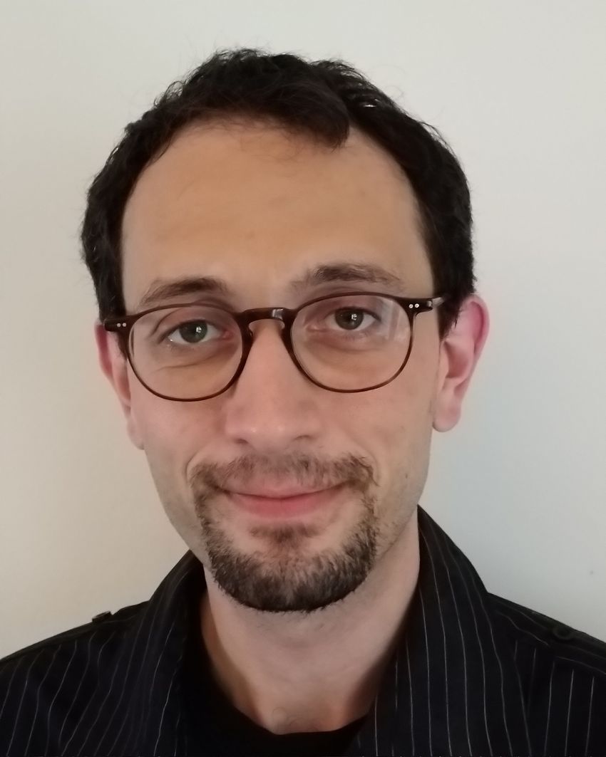 Photograph of Pieter Meysman, a computational immunologist at the University of Antwerp, Meysman has short dark hair and black-framed glasses and is standing in front of a light-colored wall. He is wearing a black collared shirt with thin stripes and is smiling at the camera.