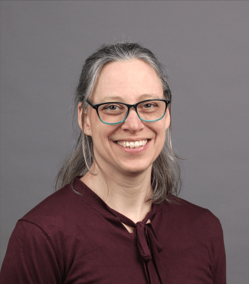 Photograph of Carmen Gerlach, a T cell immunologist at the University of Regensburg, the Karolinska Institute, and the Leibniz Institute for Immunotherapy. Gerlach is in front of a gray background and is wearing a dark red shirt and has teal and black-framed glasses. She has medium length gray and brown hair and is smiling at the camera.