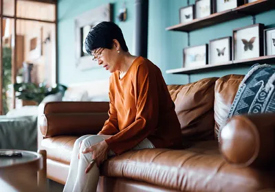 Photograph of an older woman in an orange, long-sleeved shirt sitting on a brown couch holding her knee. 
