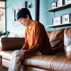 Photograph of an older woman in an orange, long-sleeved shirt sitting on a brown couch holding her knee. 
