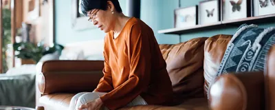 Photograph of an older woman in an orange, long-sleeved shirt sitting on a brown couch holding her knee. 