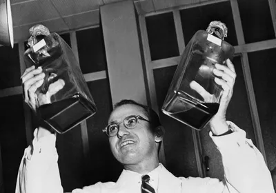 Jonas Salk wears a white lab coat and holds up two jars of liquid.