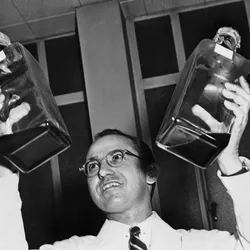 Jonas Salk wears a white lab coat and holds up two jars of liquid.