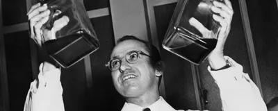 Jonas Salk wears a white lab coat and holds up two jars of liquid.