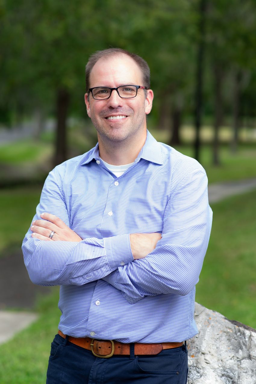 Photograph of Kyle Allen, a tissue engineer at the University of Florida, standing outside in front of a blurred background of trees. Allen is wearing black glasses and a blue collared button down shirt.