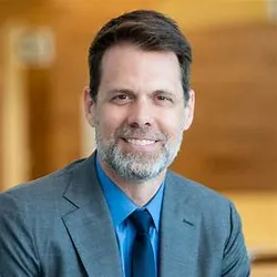 Photograph of Harold Collard. Collard has short, brown hair and a short, light-colored beard. He is wearing a grey suit jacket over a blue collared shirt and dark blue tie. Collard is smiling at the camera. 