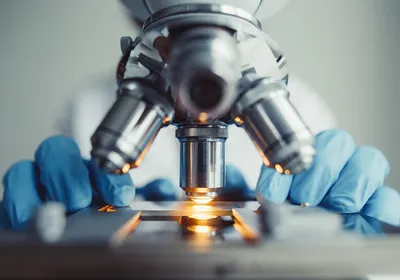 A close-up view of a light microscope with a scientist sitting behind it wearing blue gloves.