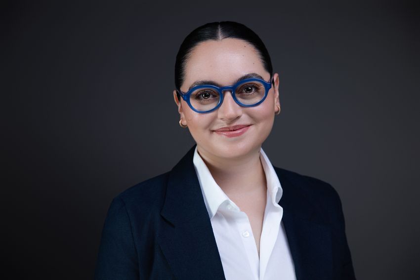 Isabella Batina wears blue-framed glasses, a dark blazer, and a white shirt in a headshot photo against a grey background.