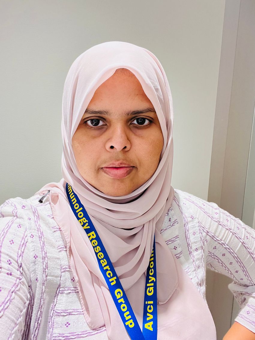 Zahra Nawaz wears a light pink headscarf, a light-colored top, and a blue conference lanyard in a headshot photo.