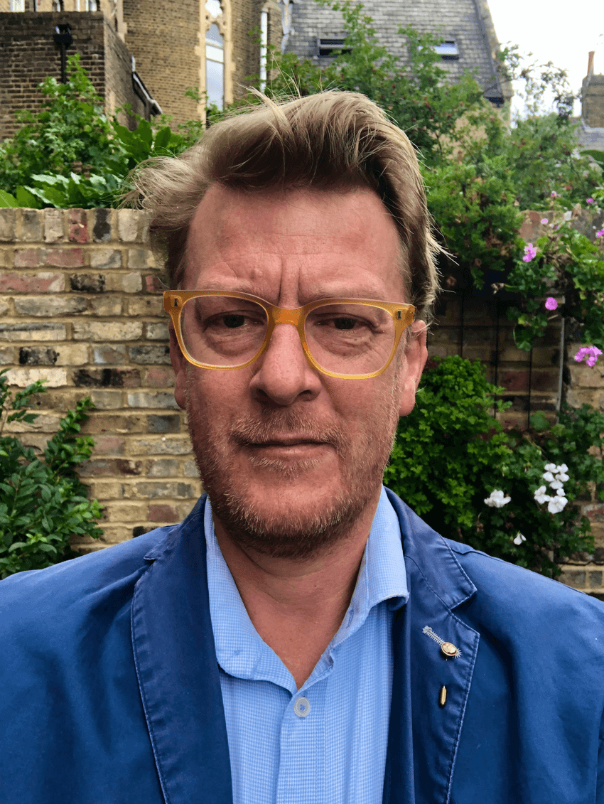Max Telford, a zoologist at the University College London, stands in front of a light brick stone wall with greenery and white and purple flowers. Telford is looking at the camera and is wearing yellow-framed glasses and a light blue collared shirt with a darker blue jacket.
