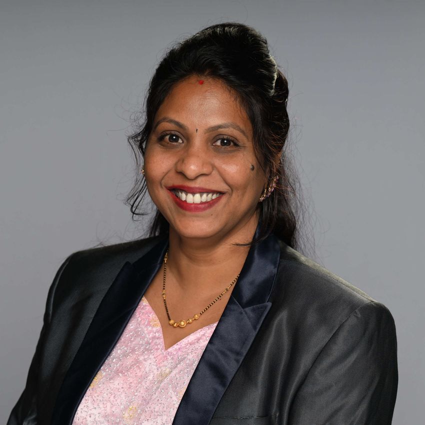 Deepika Godugu wears a dark blazer and a light shirt in a headshot photo against a light grey background.