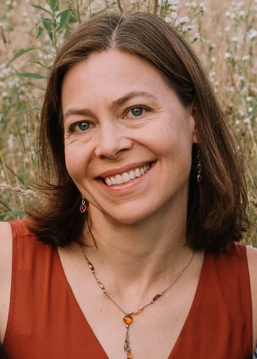 Photograph of Martha Bhattacharya, a neuroscientist at the University of Arizona, smiling at the camera in front of a blurred background of tall plants. Bhattacharya has shoulder-length brown hair and is wearing an orange sleeveless shirt with a necklace that has an orange pendant.