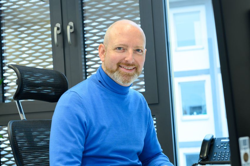 Photograph of Björn Schumacher, a biologist studying aging at the University of Cologne. Schumacher is sitting in a desk chair wearing a blue turtleneck sweater. He has a short beard and is smiling at the camera.