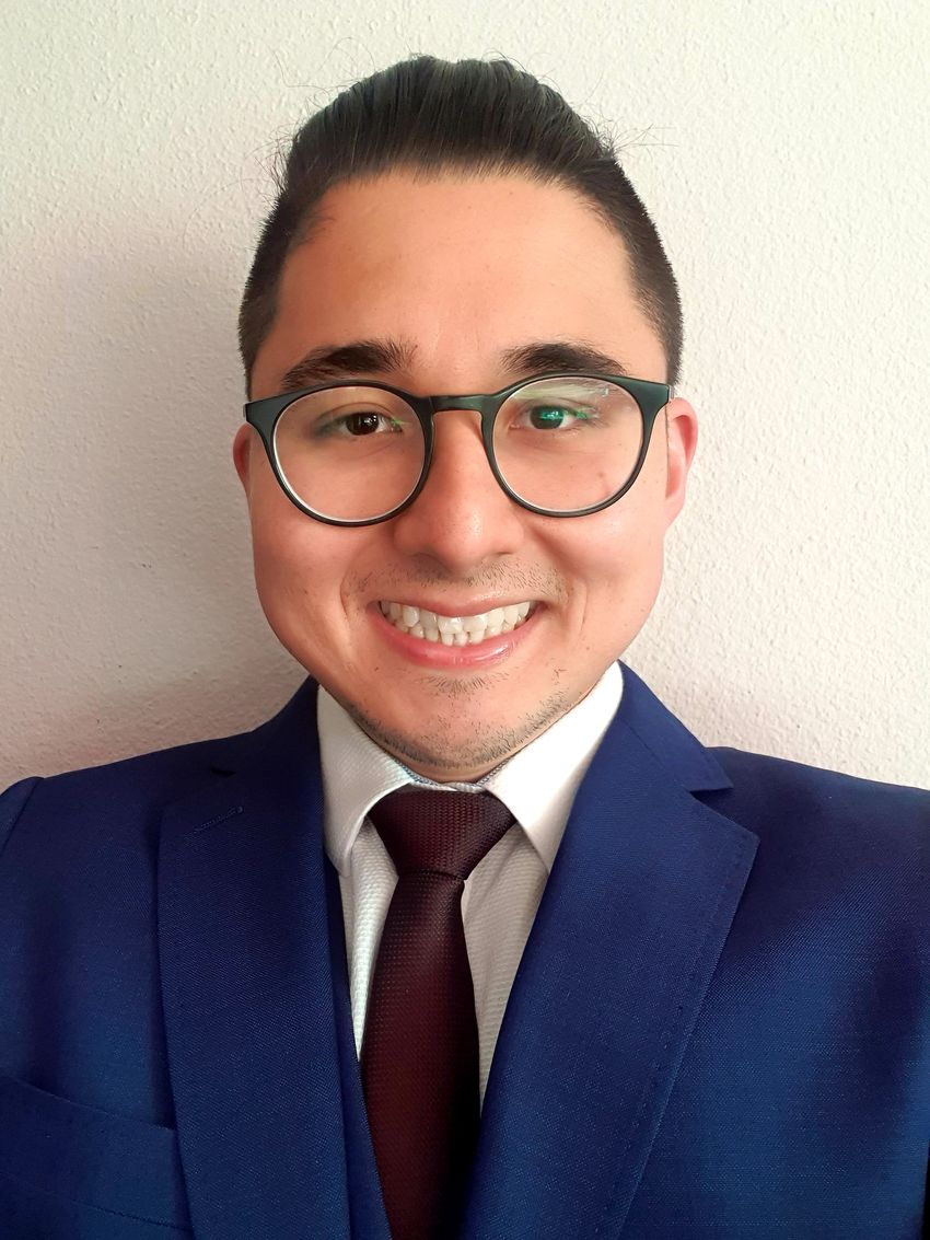 Photograph of Juliano Morimoto, an interdisciplinary researcher and biologist at the University of Aberdeen, is smiling at the camera standing in front of a light colored background. He is wearing a dark blue suit jacket, a white collared shirt, and a dark tie. He has short dark hair and circle-framed glasses.