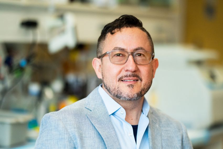 Photograph of Joseph Arboleda-Velasquez, a geneticist at Massachusetts Eye and Ear and Harvard Medical School. Arboleda-Velasquez is standing in front of a blurred background of a lab and is wearing a light gray suit jacket over a light blue collared shirt. He has short dark hair and glasses.