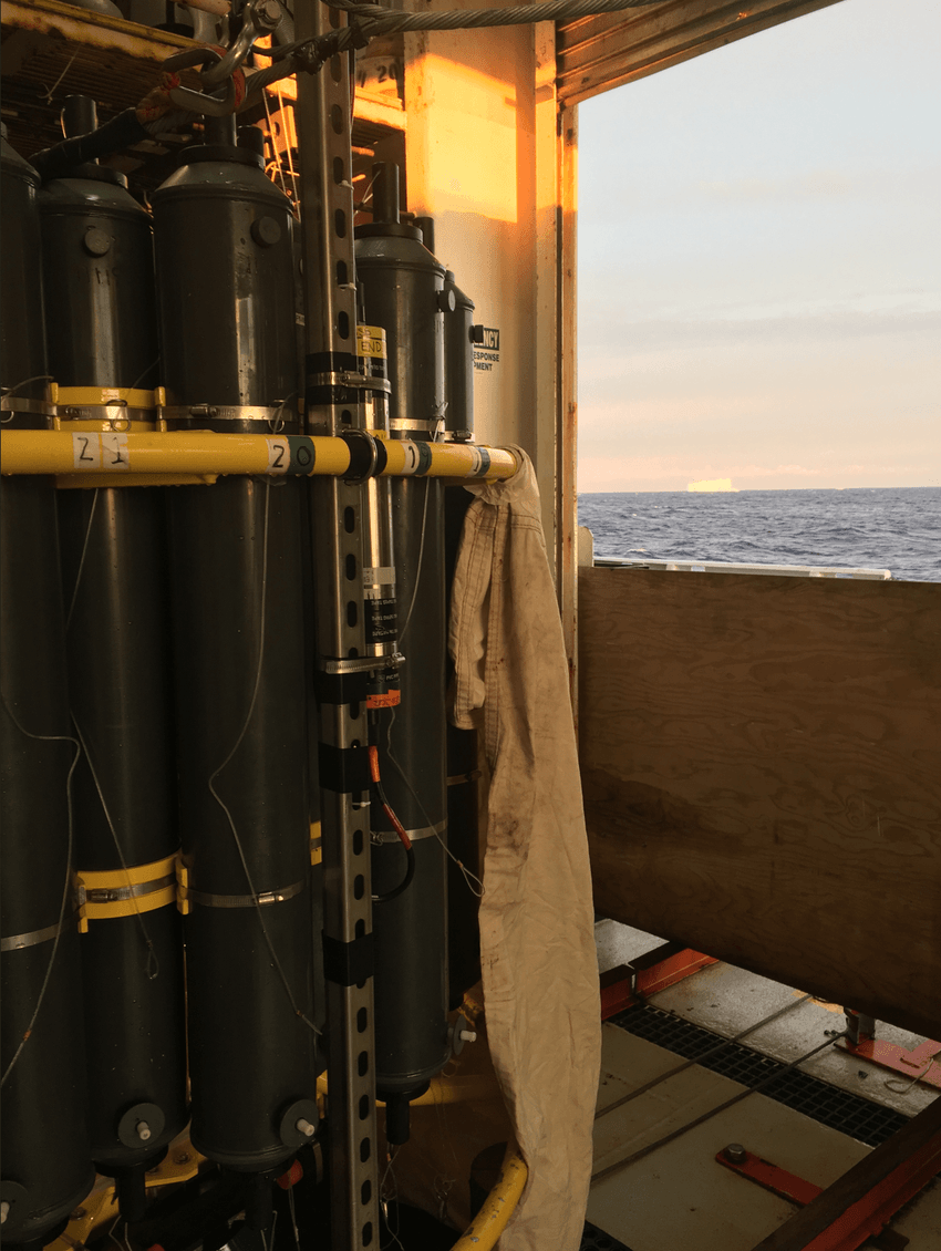 A group of 20-liter black water collection tubes stand against a wall with a view of the ocean and an iceberg in the distance.