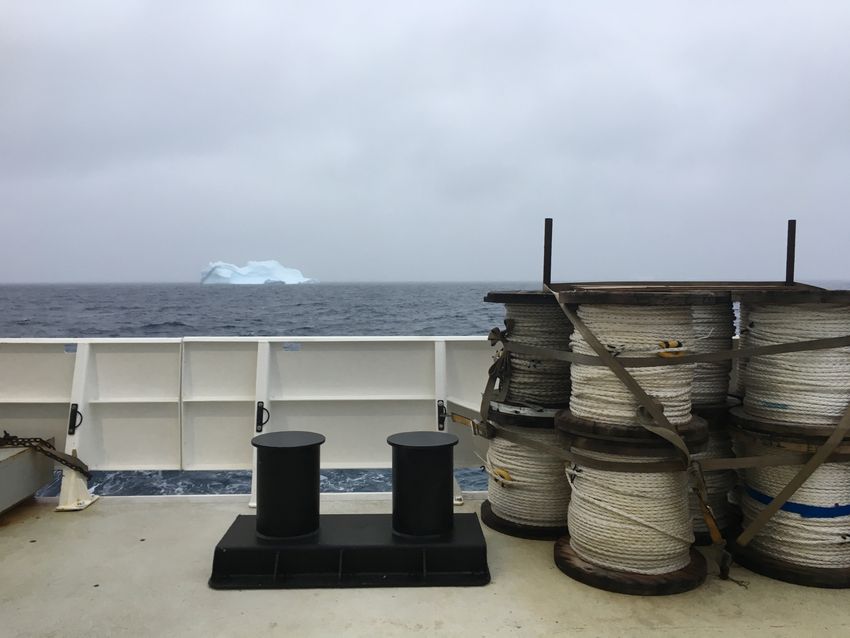 Giant spools of cables sit on the deck of the boat, and the ocean with an iceberg is seen in the distance.