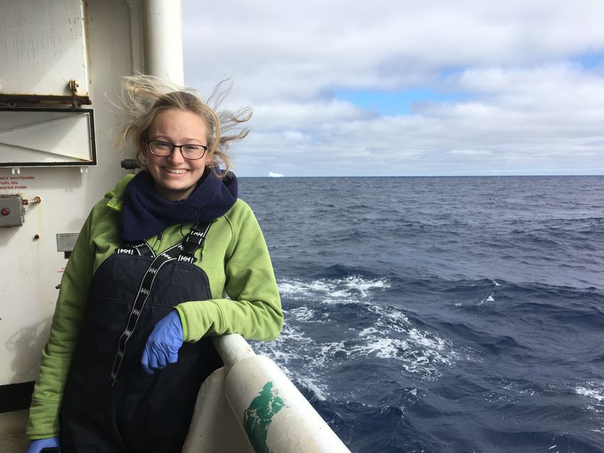 Bethany Kolody wears a green jacket, black scarf, and blue gloves as she leans against an open window looking out into the ocean with an iceberg floating in the distance.
