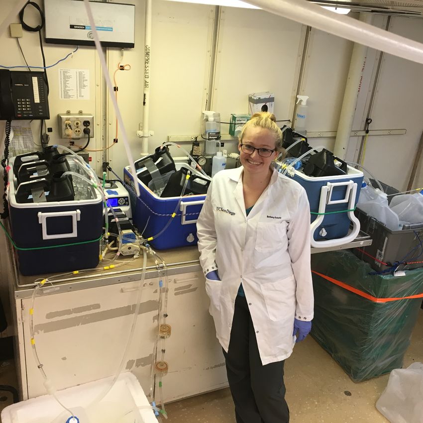 Bethany Kolody wears a white lab coat while standing in front of her water filtering set up on the GO-SHIP research vessel.