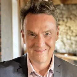 Derek Bowie wears a grey suit jacket and a collard shirt in a headshot image.