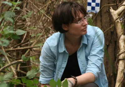 Kirsty Wright is searching for evidence in a missing persons investigation. She is wearing a blue shirt and glasses. Behind her is dense vegetation and police procedural tape. 