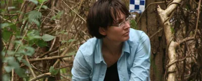 Kirsty Wright is searching for evidence in a missing persons investigation. She is wearing a blue shirt and glasses. Behind her is dense vegetation and police procedural tape. 