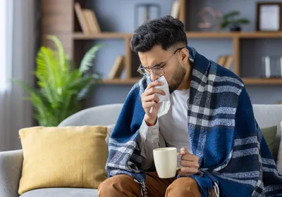 Image of a young man sitting on a sofa. He has a blanket wrapped around his shoulders. He has a tissue raised to his face while he holds hot beverage in his other hand.