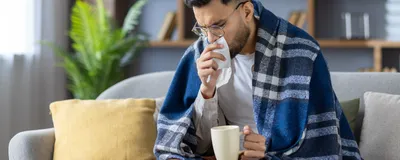 Image of a young man sitting on a sofa. He has a blanket wrapped around his shoulders. He has a tissue raised to his face while he holds hot beverage in his other hand.