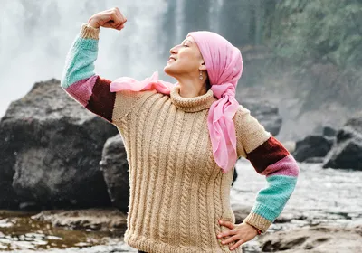 A female patient with cancer, wearing a multicolored knit sweater and a pink headwrap, poses in front of a waterfall. She is flexing her right arm, symbolizing strength and victory in the fight against cancer.