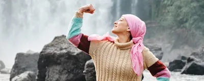 A female patient with cancer, wearing a multicolored knit sweater and a pink headwrap, poses in front of a waterfall. She is flexing her right arm, symbolizing strength and victory in the fight against cancer.