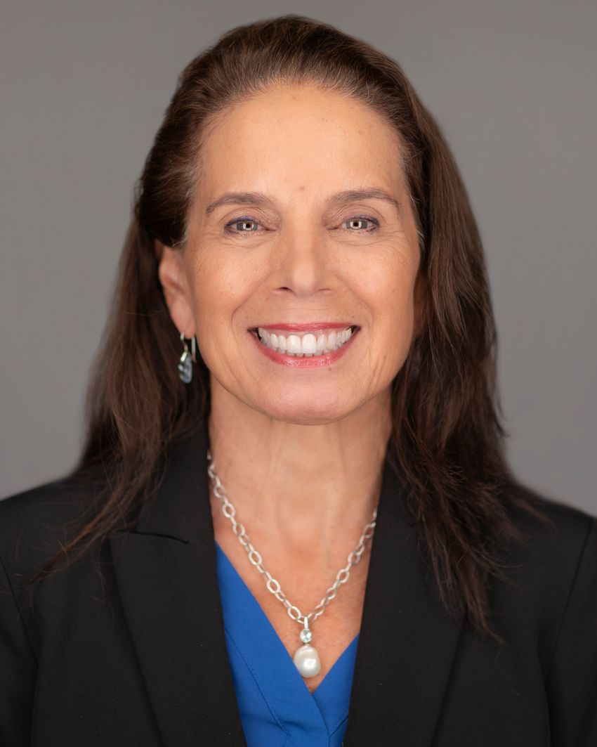 A headshot of Anna Giuliano wearing a blue top and black coat. She wears a pearl necklace and earrings, and smiles at the camera.